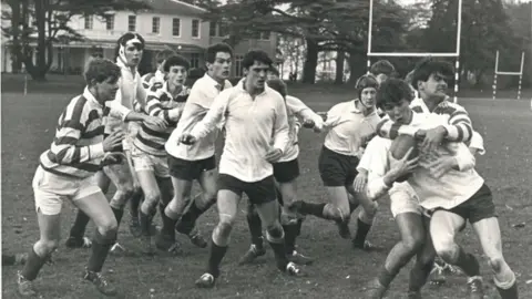 Framlingham College Black and white image of a group of men, some in white polo shirts and some in striped polo shirts, playing rugby union. One player has the rugby ball and the others are looking on as another player tackles him. 