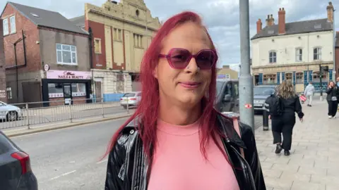 Elizabeth Baines / BBC A woman with red hair wears pink sunglasses. She wears a pink top and black leather jacket. Behind her a highstreet is blurred.