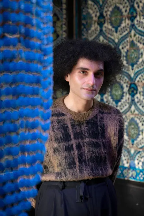 RBKC/ Jaron James A man with big black hair looks at the camera. He is wearing black slacks and a patterned brown and black sweater