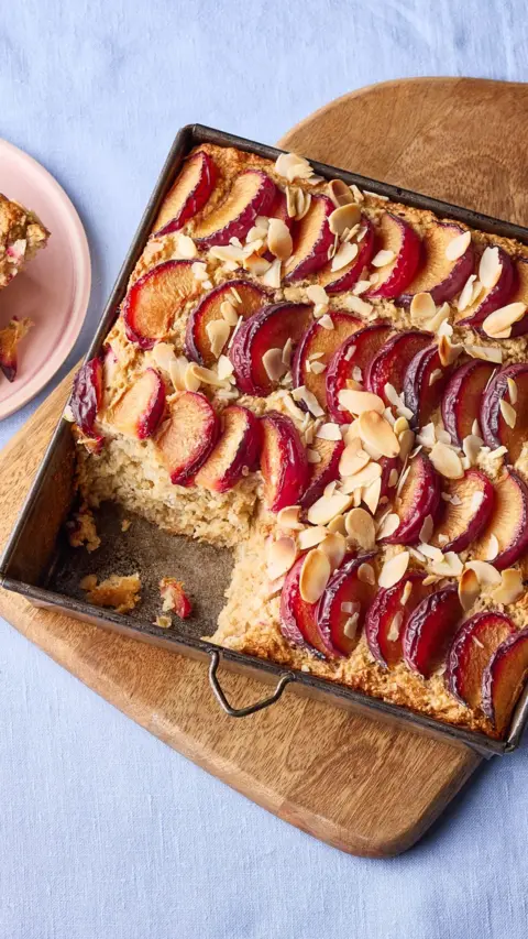 A baking tray containing a plum and almond bake, sitting on a wooden board. A slice has been removed.