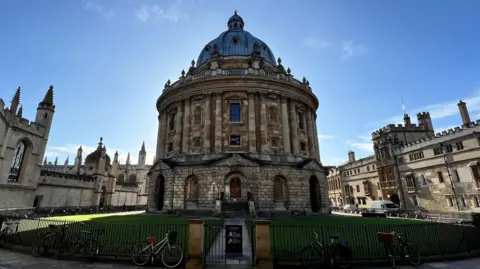 The Radcliffe Camera on a sunny day. The building itself is in shadow and is backlit against a blue sky. Other historical buildings can be seen around it, with a round lawn and cobbled street between them.