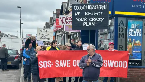 A group of people walking down the street holding placards and signs opposing the Portswood bus gate scheme.