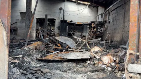 The inside of a workshop is completely burnt, with twisted metal and black and brown charring everywhere.