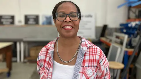 A woman with dark hair tied up, wearing glasses, a silver chain and a white top with a checked shirt over it. 
