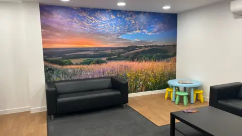 A view of a room with wood laminate floors and a dark grey rug. There are two black boxy leather sofas and a black coffee table. There is also a small light blue table and four chairs for children. On the wall at the back of the photograph is a landscape photo of a countryside scene at sunset