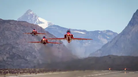 The Red Arrows taking off from Greenland on a previous Atlantic crossing. There are snow-capped mountains in the background and a runway beneath the aircraft.
