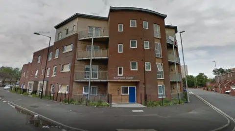 A four-storey block of flats built in brown bricks on the corner of a residential road. The entrance is curved, with balconies on either side of the central pillar.