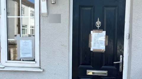 A black front door has an A4 sheet of paper full of text sellotaped to the middle of it. There is a similar notice on the window to the left of the door.