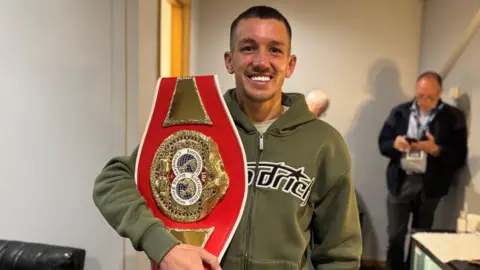 A man with short brown hair is smiling and wearing a green zip hoodie. He is holding a large red boxing belt trophy that has gold accents and two circles on it