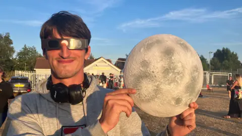 Kian Lodge smiling at the camera, holding a toy Moon. He is wearing black eclipse glasses, black headphones around his neck and a grey hoodie. He is standing in a field. The sky is blue.