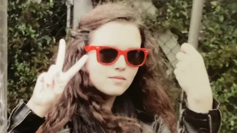 PA Media Maeve with red rimmed sunglasses on, her brown hair is partially tied back and she is doing a peace sign with one hand and holding what appears to be a swing with the other