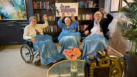 Sutton Rose Care Home Left Maureen , Ann in the middle and Doreen on the right. Three elderly women sat in wheelchairs with blue blankets over them. They have hands free microphones on. Behind them there is a book shelf with a sign that reads 'Do you remember the time?'. They have their hands in the air and they are posing for the cameras. 