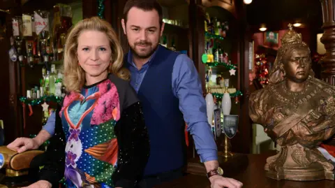 BBC/Kieron McCarron A bust of Queen Victoria is on the bar of the Queen Vic in EastEnders. Cast members Linda and Mick Carter are stood behind the bar, smiling at the camera