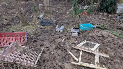 RSPCA Debris strewn across the site, including broken plastic trays and wood. There is thick mud on the ground.