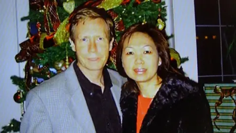 A police handout of Peter Chadwick and his wife Quee Choo Chadwick. They are shoulder to shoulder and looking at the camera, stood in front of a Christmas tree. He is in a grey suit jacket and black shirt, she is wearing a red top and black jacket.