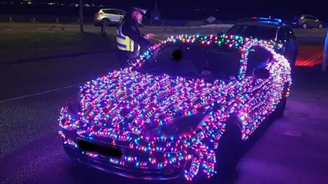 Poole Police Car covered in coloured Christmas lights stopped at the side of the road with a police car behind it and an officer standing at the driver's door.