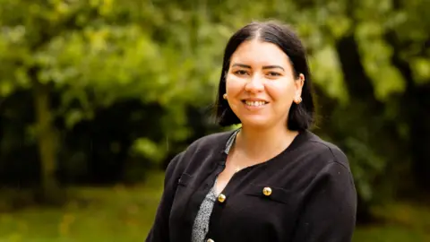 Society Without Abuse Louisa Wrighton smiles at the camera. She is wearing a black cardigan with gold buttons. Blurred behind her is some greenery.