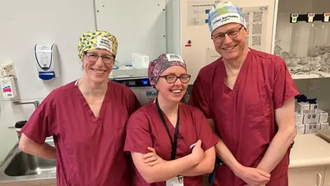 Clinicians Ingrid Wilkins, Mhairi Stewart and Ludger Barthelmes, wearing their brightly patterned reusable hats. They have matching burgundy scrubs and stand together in a hospital preparation area.

