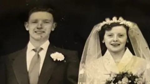 A couple on their wedding day. The man is wearing a suit and the woman wears a white dress and carries flowers.