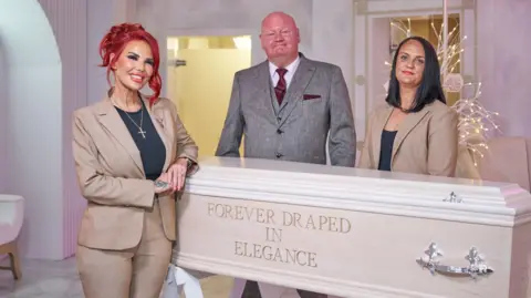 Hayley McCaughran, who has dyed red hair tied back and is wearing a light brown suit jacket and trousers over a black top, stands next to a light wooden coffin with the words 'Forever Draped in Elegance' engraved into the side. Next to her are her colleague, a bald man in a light grey suit, and a woman with black hair and wearing a similar suit to McCaughran. 
