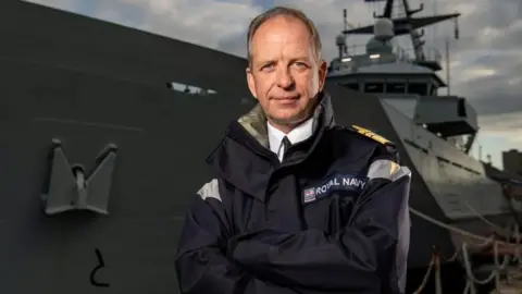 Vice Admiral Jeremy Kyd is standing in front of a naval vessel. He is wearing Royal Navy uniform with epaulettes and has his arms crossed. He is smiling and has short grey hair and blue eyes.