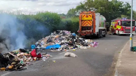 A large pile of discarded waste spread across a road, with smoke rising from a section of the rubbish in the foreground. A refuse collection vehicle and a fire engine are positioned nearby. Dense hedgerows line one side of the road, while a pavement and streetlight are visible on the opposite side. 