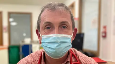 Consultant geriatrician Rhodri Edwards has short grey hair and is wearing a red and white chequered shirt and a blue face mask. 