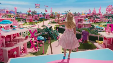 Warner Bros A Barbie doll dressed in a pink chequered dress looks out at a Barbie pink paradise in a scene from the Barbie movie.