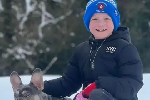 Joshua Slater pictured with a pug dog in the snow. Joshua is wearing a black jacket and a blue hat and is smiling in the photo.