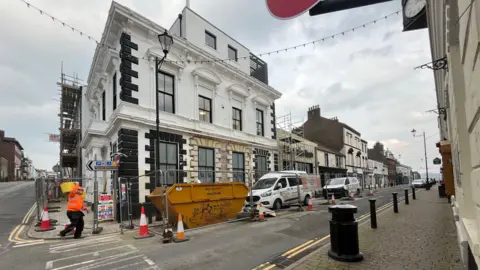 A general view of the Carlton cinema taken from the corner of the street, looking up Senhouse Street. The building is still being refurbished and there is scaffolding and metal fencing around the building. There is a workman in orange high viz clothing carrying something to a skip outside the building, next to a van parked outside. The building is made of two main section. The left side is an 1830s three-storey building painted white with some black detailing, including the sash windows. Around the windows are some decorations such as arches. At the top of the building there is a roof terrace surrounded by a black fence. The section on the right is a metal sage green building on one level, with a tall pitch roof. It has a large window and entrance at the front and the name of the building across the top.