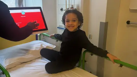 DKMS A young boy sits on a hospital bed. He is wearing dark coloured pyjama clothing. Beside him is a monitor and a hand touching the screen. The screen is showing a red and white display.