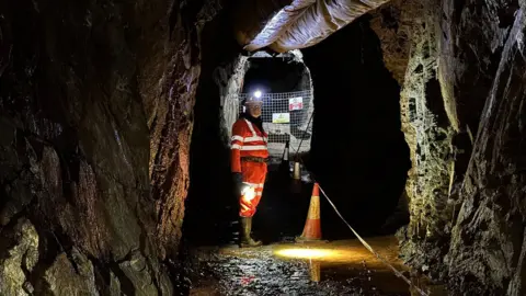 BBC A mine worker in orange high viz clothing, a hard hat with a lamp on it and boots stands in a mine tunnel with dark grey walls and water on the ground 