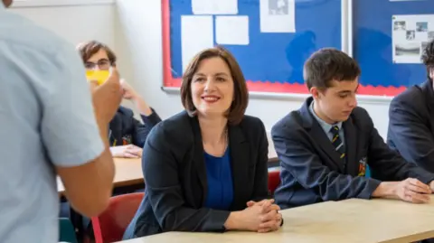 Phillipson wears a black suit jacket over a royal blue dress with a silver necklace. Her hair is worn in a dark brown, chin-length bob. She wears bright red lipstick and is smiling at a teacher, who can partially be seen in the foreground on the left. There are pupils sitting around her, all wearing a navy school uniform with a pastel blue shirt and a navy tie with pale blue and gold diagonal stripes. 