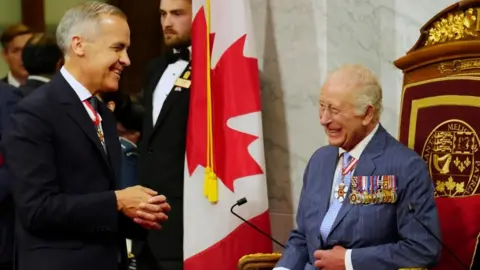 Mark Carney, smiling with his hands clasped in front of him, stands facing King Charles, who is seated and smiling.