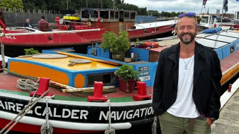 The boat is brightly coloured and has plant pots on it. Other tugs are in the background. John stands at the side of the boat smiling with his hands in his pockets and his sunglasses on his head.