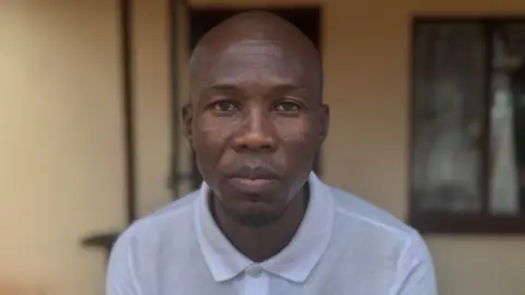 A black man in a white polo shirt looks into the camera.