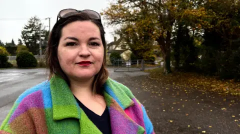 A woman in a head and shoulders image. She is wearing a brightly-coloured coat in purples, greens, blues and pinks, and a black top underneath. She has glasses perched on top of her head. She has dark brown eyes and dark brown hair. She is standing on a path beside some trees. The ground behind her is wet, with leaves on the ground. 