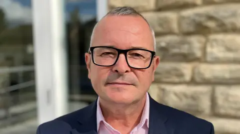 Lee Barron with short white hair and black-framed glasses, wearing a blue jacket and pink shirt standing in front of a stone wall