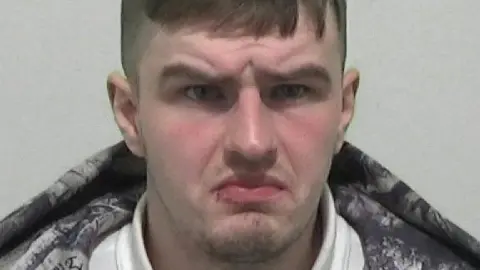 Northumbria Police Mugshot of Daniel Gaunt. He has a faint goatee beard and short brown hair. His brow is furrowed and he looks confused. 
