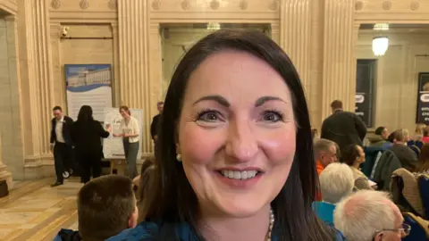 A woman with dark hair, a pearl necklace and blue dress is smiling at the camera. Behind her is a room full of people