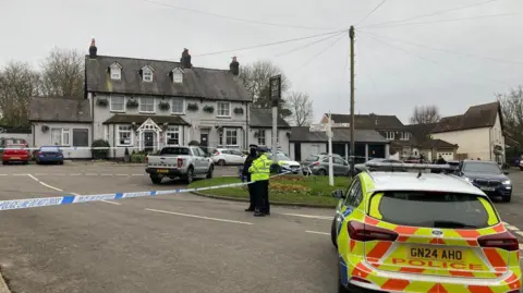 Dan MacLaren/BBC Police stand outside the pub, which is taped off. A police car is in the foreground.