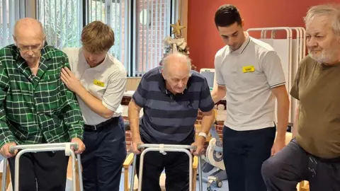 NHFT A group a people, three older men, and two workers, carrying out rehabilitation work. They are in a room, with red walls, and they are all looking down.