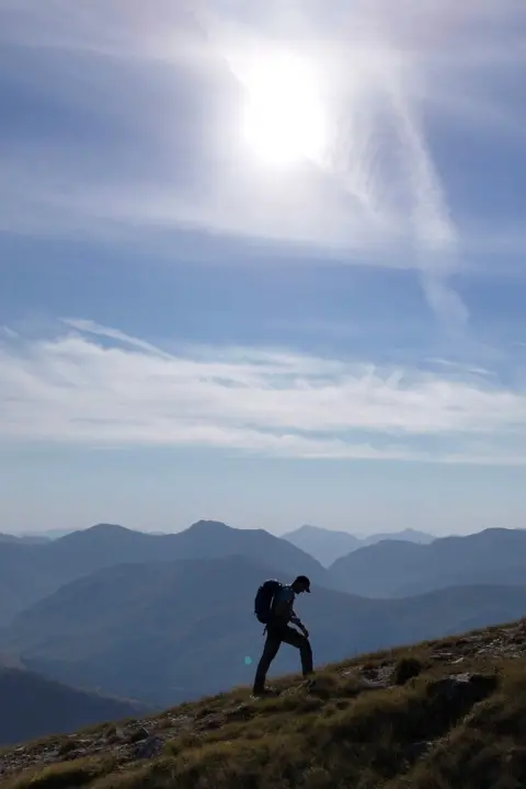 Kyle Lenaghen  A hillwalker ascending a slope against a backdrop of mountains