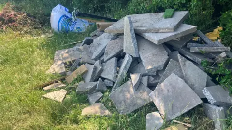 BBC / Elizabeth Baines A pile of concrete flags. The flags are broken and sit beside builders waste on green space.