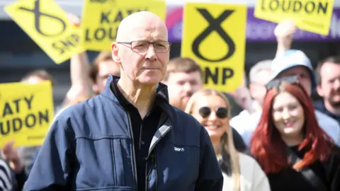 John Swinney - a bald man with glasses and wearing a dark blue jacket with a small SNP logo on it, stands in front of a group of supporters waving SNP placards 