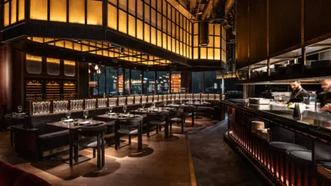 Gordon Ramsay Restaurant Global A still of Gordon Ramseys Lucky Cat restaurant, theres a row of tables under a quirky yellow light panel. The tables are lined with wine glasses and cutlery, with chefs working in the open kitchen beind. 