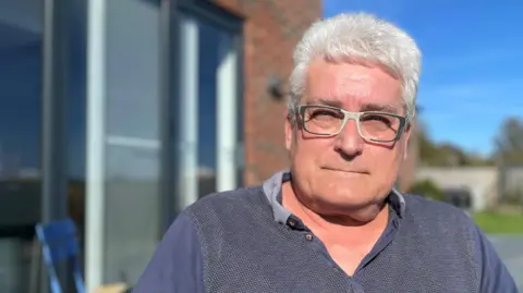 BBC Mike Crookes sits in a garden outside a house on a sunny day. He has bushy, white hair and wears glasses with black and white frames, and a grey and blue polo shirt.