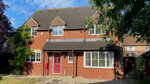 The front of a family home with a red door and a porch type area over the front door. It has three upper windows and three downstairs. There are trees in front of the property and a some grass.