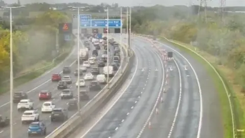 Highways England CCTV still image of traffic jams on one side of a motorway and four empty lanes on the other side. It appears to be raining.