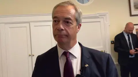 A man looking forwards and to the side of the camera. He is wearing a dark suit and dark purple tie and white shirt. Behind him are white cupboards and another man who is wearing a suit is looking down. 